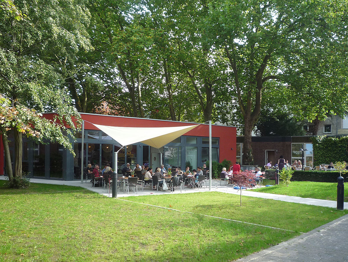 Ein Foto vom Café des Alexianer Krankenhauses in Aachen mit Menschen und viel Grün von außen fotografiert